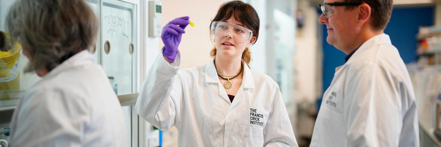 Three people wear lab coats and talk to each other in a lab