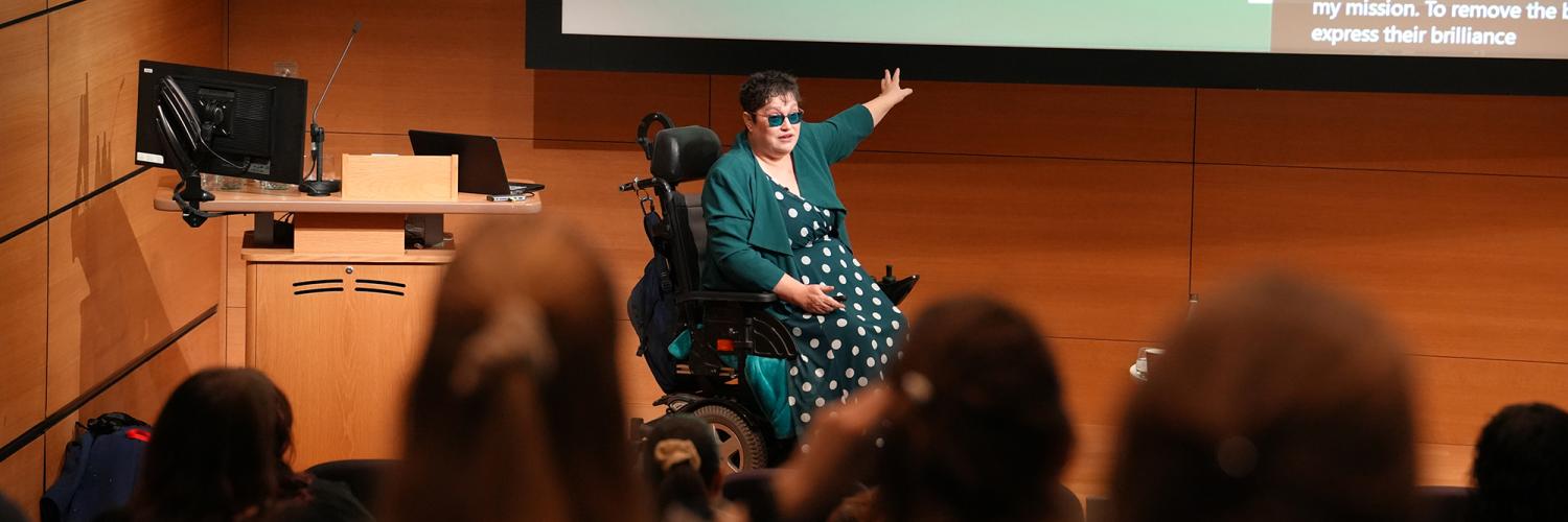 A women who uses a wheelchair, wearing glasses and a green dress with white polka dots, gestures towards the screen as she gives a presentation in the auditorium