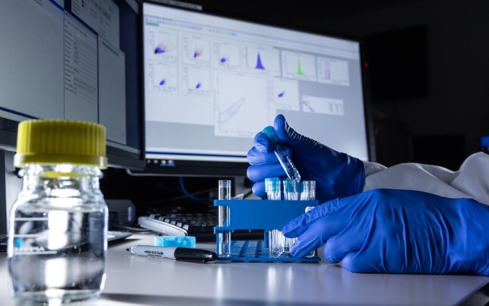 Hands wearing gloves holding a pipette with a computer screen in the background