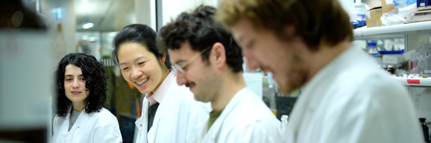 Four people in lab coats are in a line and smiling to each other as they look down at a workbench