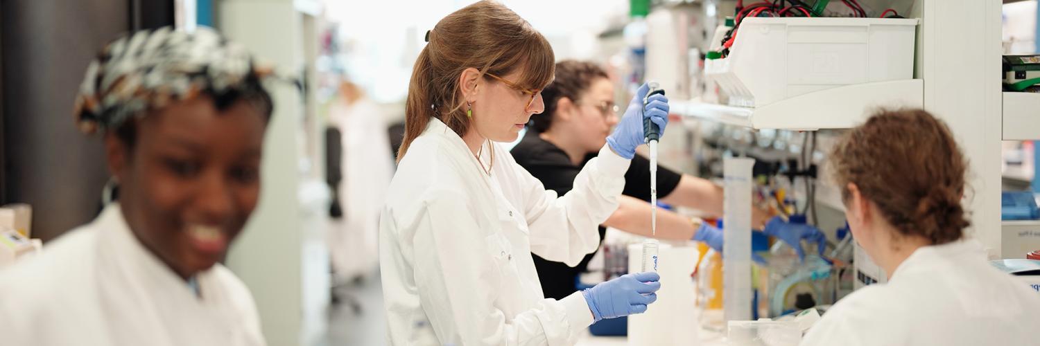 a group of researchers working in a lab, some are at the lab bench using a pipette, others are walking in the foreground