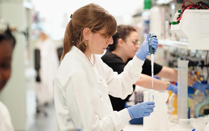 a group of researchers working in a lab, some are at the lab bench using a pipette, others are walking in the foreground
