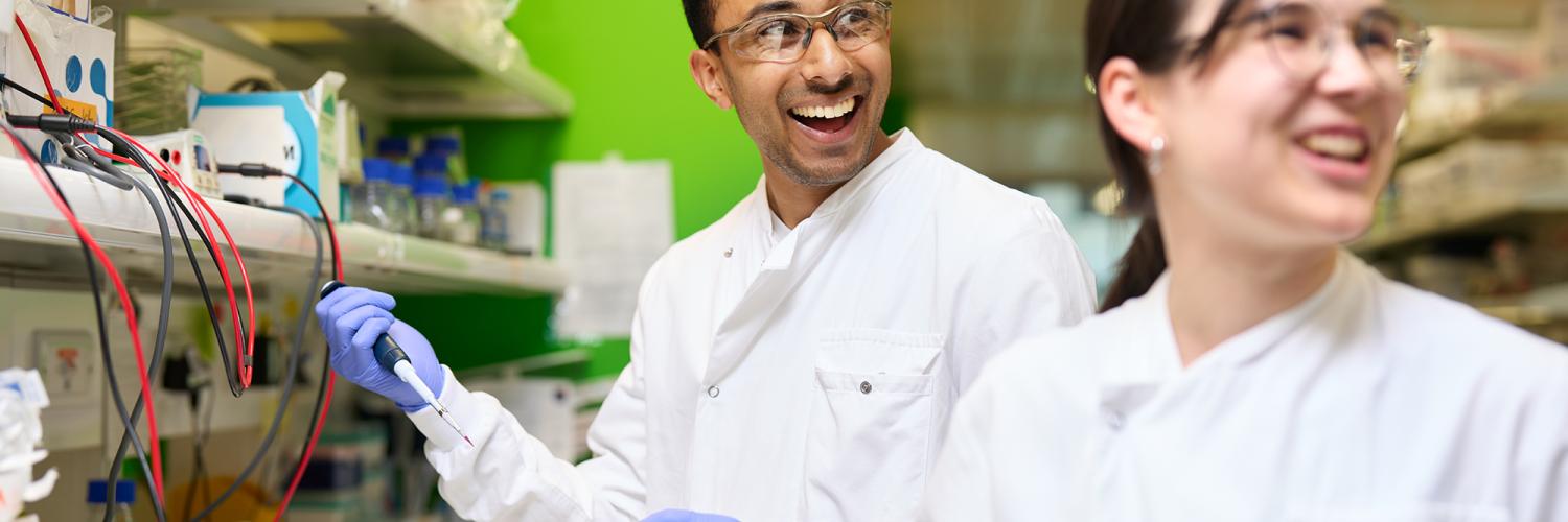 Two researchers in white lab coats smile at someone behind them and out of the frame, one of them is holding a pipette